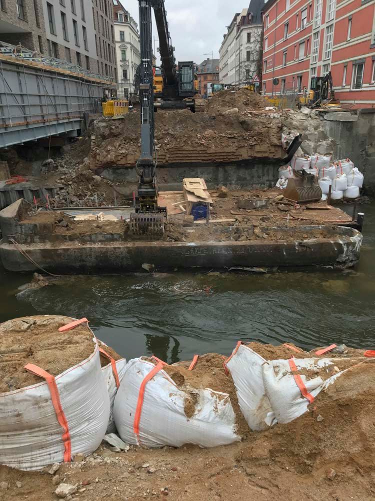 Baustelle an einem Fluss mit einem Bagger, der Erde und Schutt wegräumt. Weiße Sandsäcke im Vordergrund.