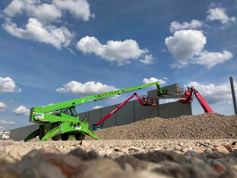 Zwei Bauaufzüge in der Nähe einer Betonmauer und eines Steinhaufens unter einem teilweise bewölkten blauen Himmel.
