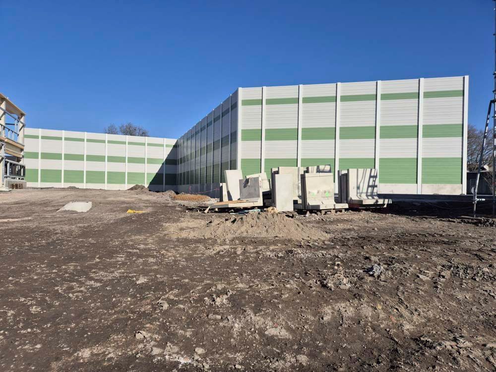 Eine lange, weiß-grüne Lärmschutzwand im Bau auf einer Baustelle unter klarem, blauem Himmel.