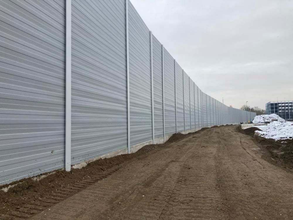 Ein langer, grauer Lärmschutzzaun neben einer unbefestigten Straße unter einem bewölkten Himmel.