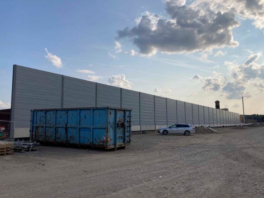 Blauer Müllcontainer, silbernes Auto und hohe graue Schallschutzwand auf einem Schotterplatz unter einem teilweise bewölkten Himmel.