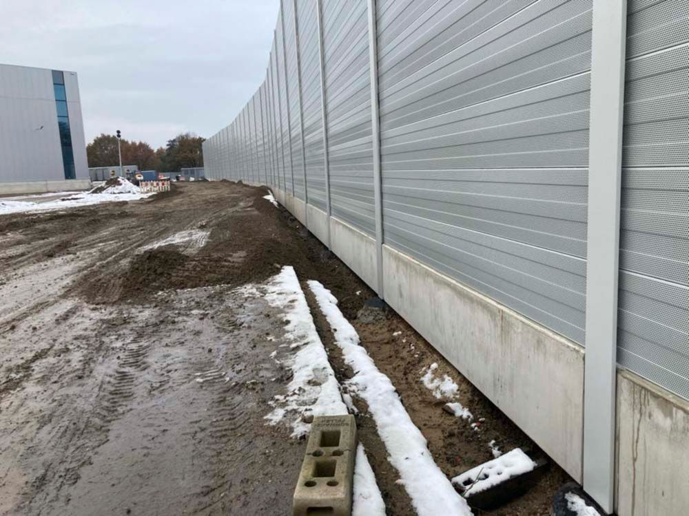 Eine lange graue Lärmschutzwand neben schlammigem Boden mit Bauschutt und einem Gebäude im Hintergrund.