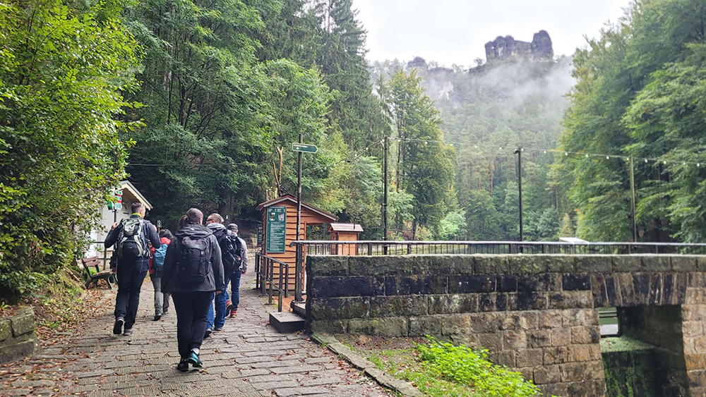 Group of people walking on a stone path along a river; forested area with a misty rock formation in the background.