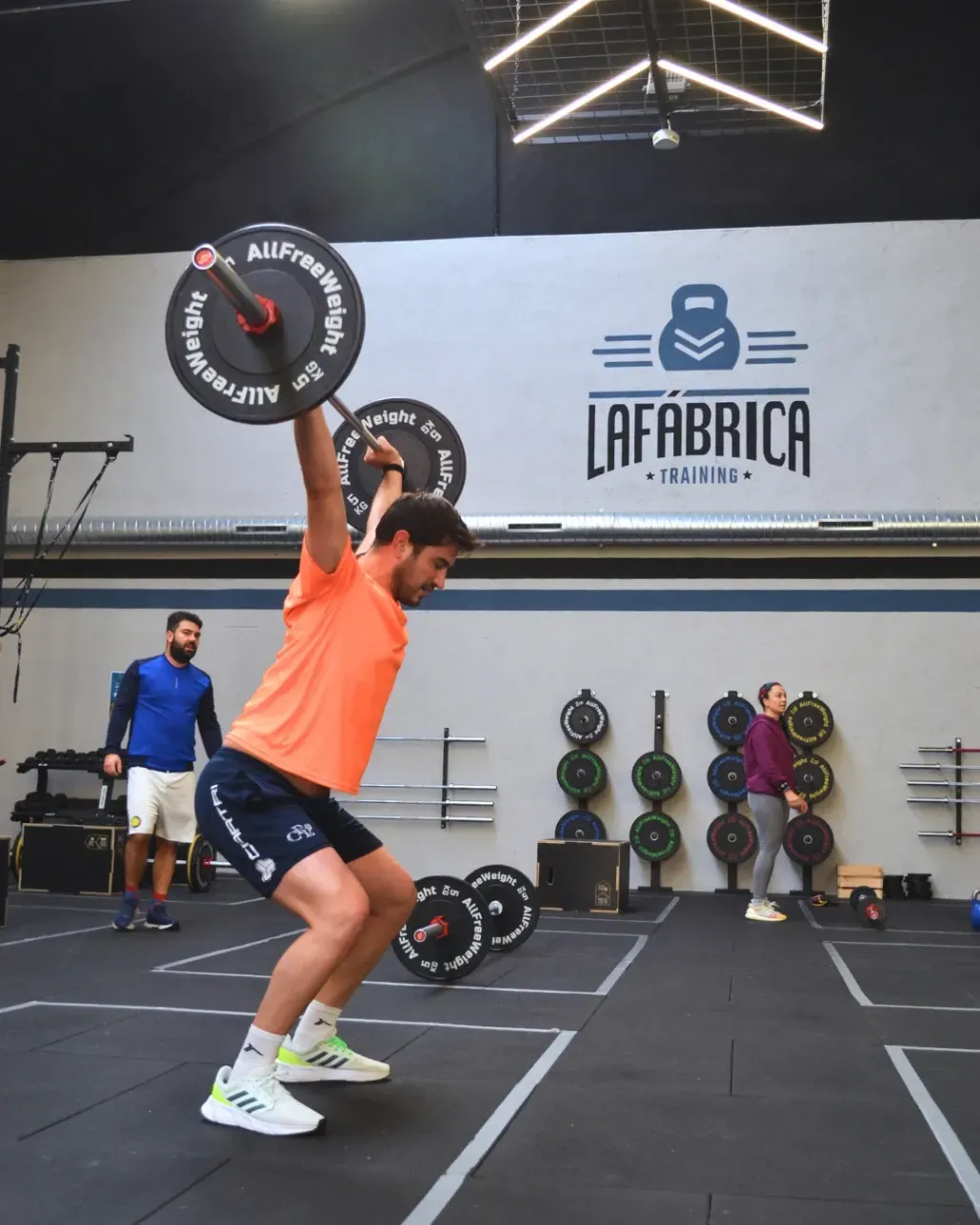 Un hombre levanta una barra por encima de la cabeza en el gimnasio mientras otros observan.