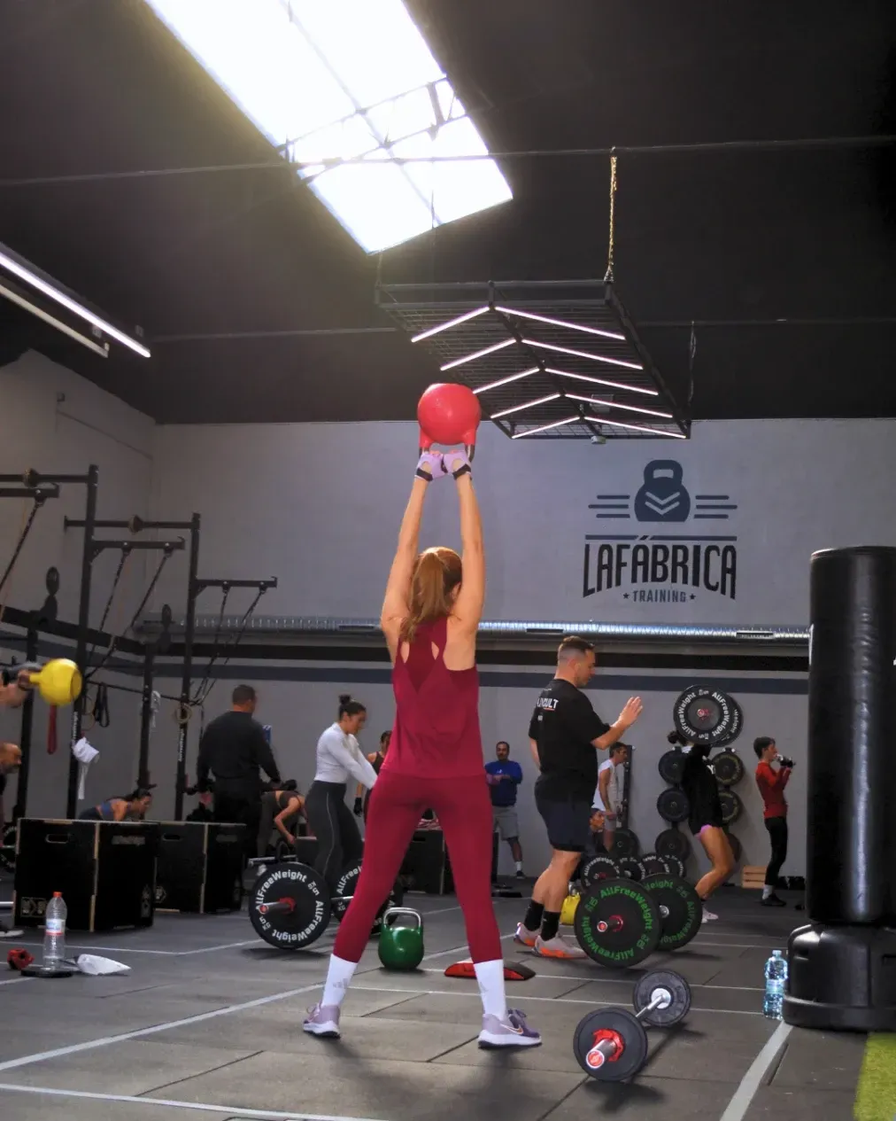 Una mujer con traje rojo levanta un balón medicinal en un gimnasio. Otras personas hacen ejercicio al fondo.