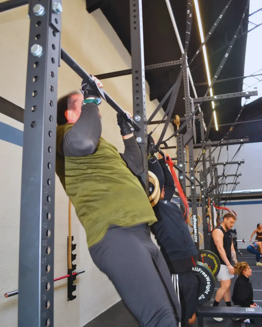 Una persona hace una dominada en una barra negra en un gimnasio. Otras personas entrenan al fondo.