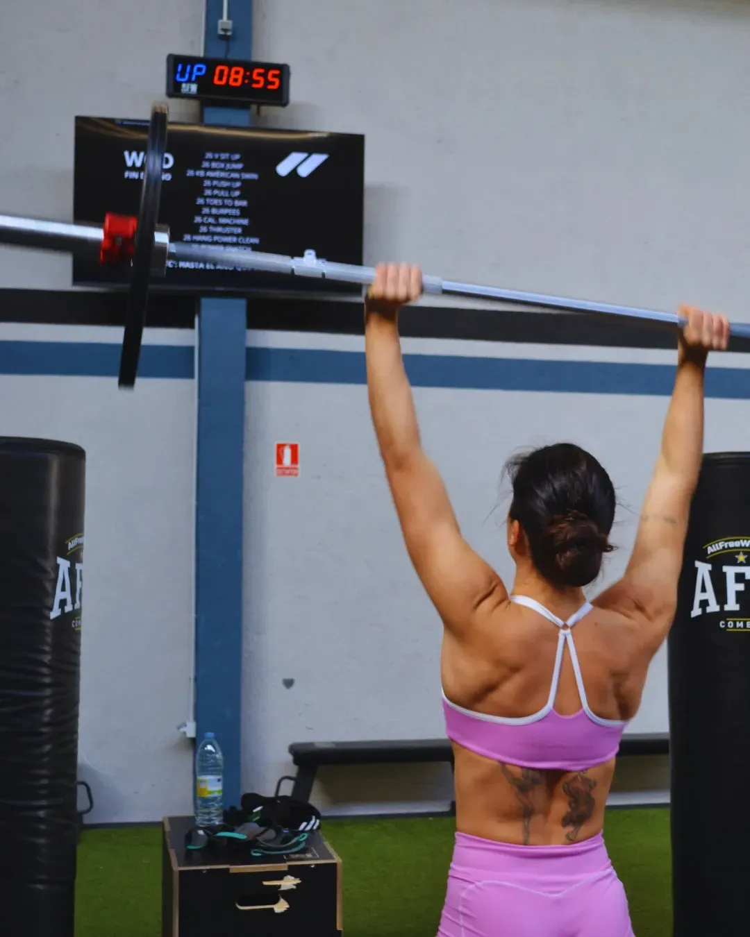 Mujer levantando pesas en el gimnasio. Ropa deportiva rosa, brazos extendidos. Saco de boxeo negro, cronómetro a la vista.