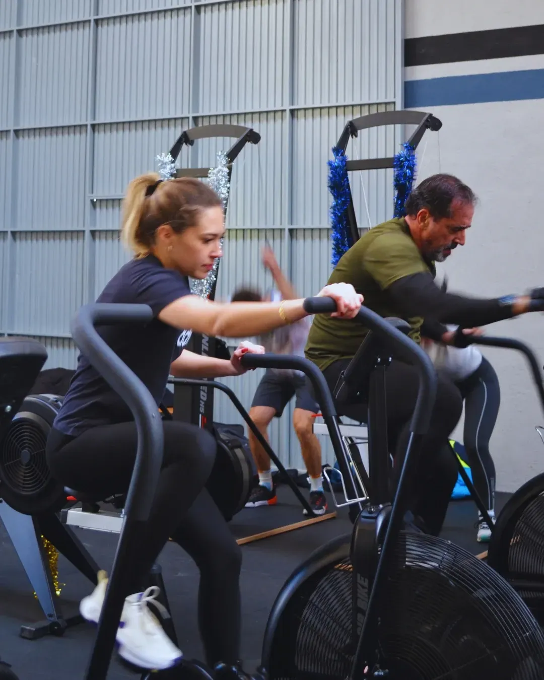 Personas en bicicletas estáticas en un gimnasio. Una mujer con leggings negros y un hombre con camisa verde. Otros al fondo.