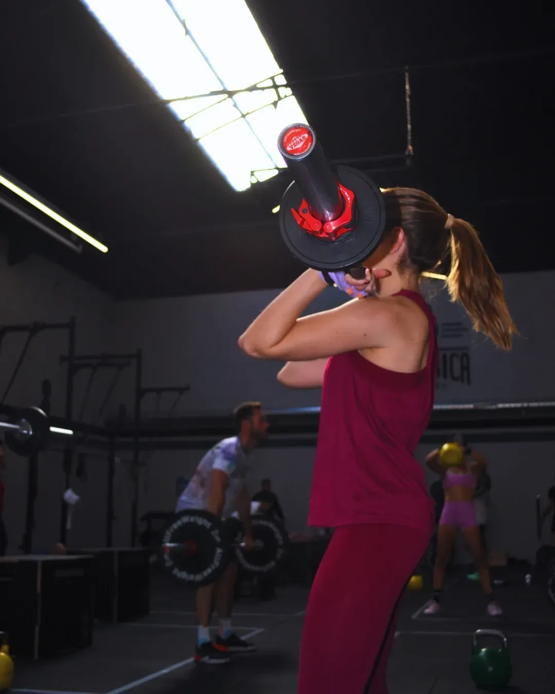 Mujer con ropa deportiva de color burdeos haciendo un ejercicio de hombros con una barra con pesas en un gimnasio.