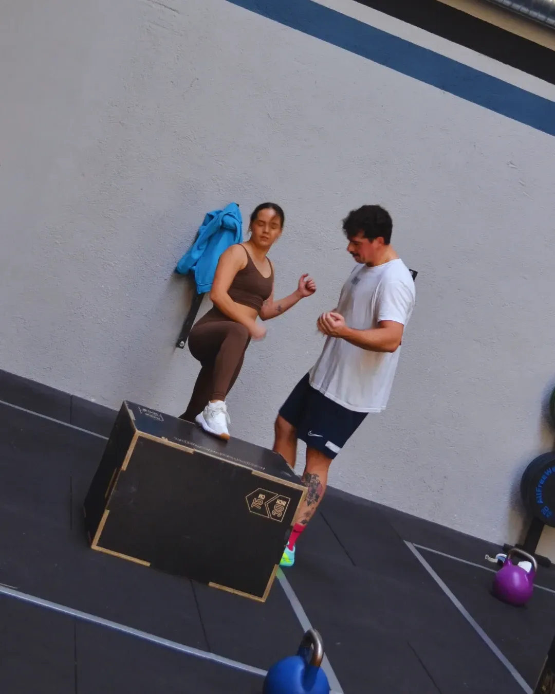 Mujer en la caja, hombre observando; ambos con ropa deportiva. Gimnasio en tonos grises y marrones.