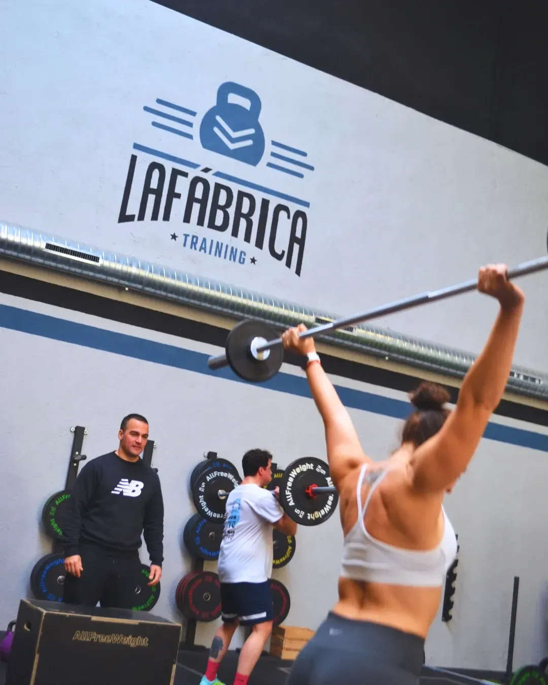 Mujer levantando una barra en el gimnasio; entrenador y observador observan. Logotipo de 