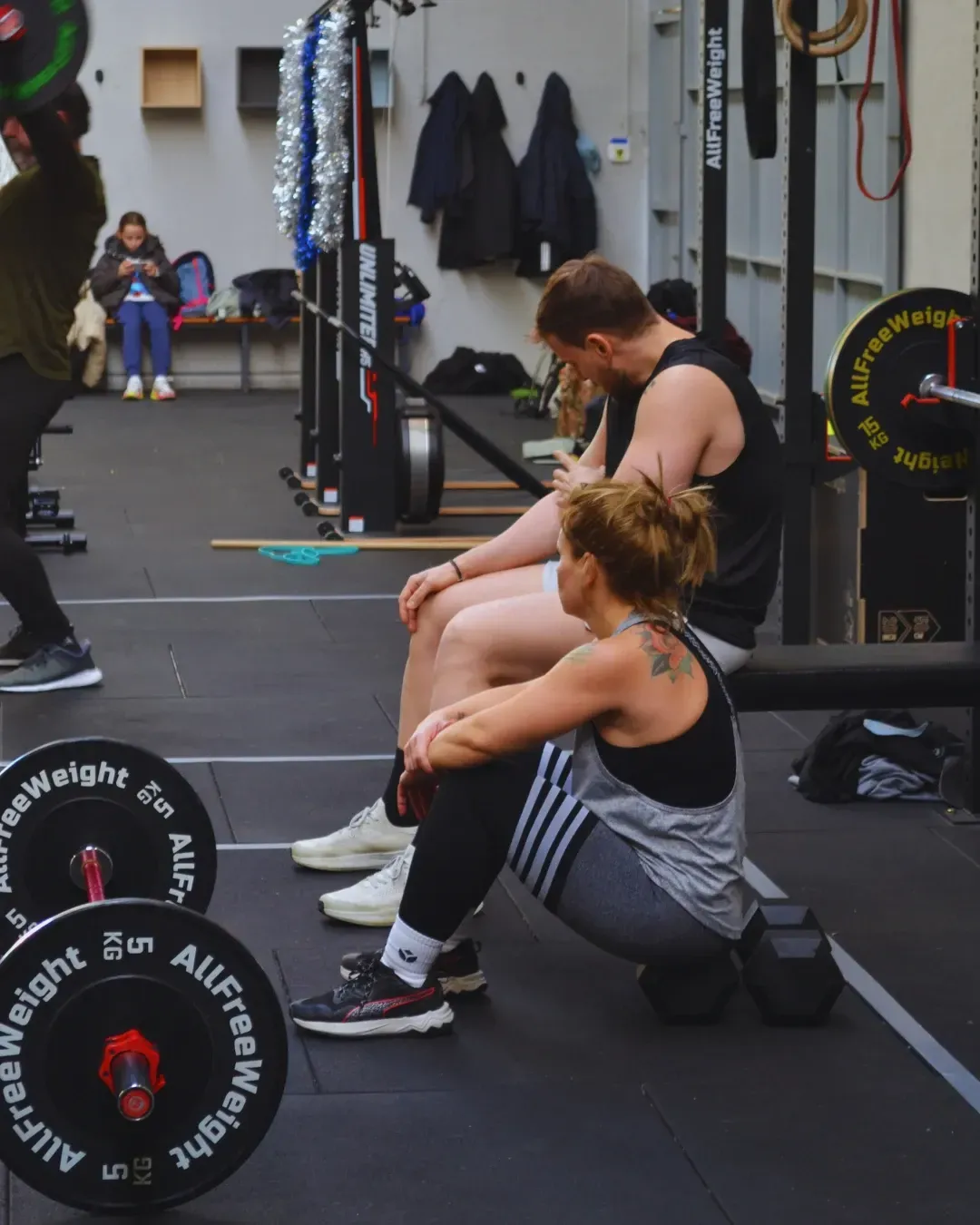 Personas descansando en un gimnasio después de entrenar. Un hombre y una mujer sentados junto a una mancuerna.