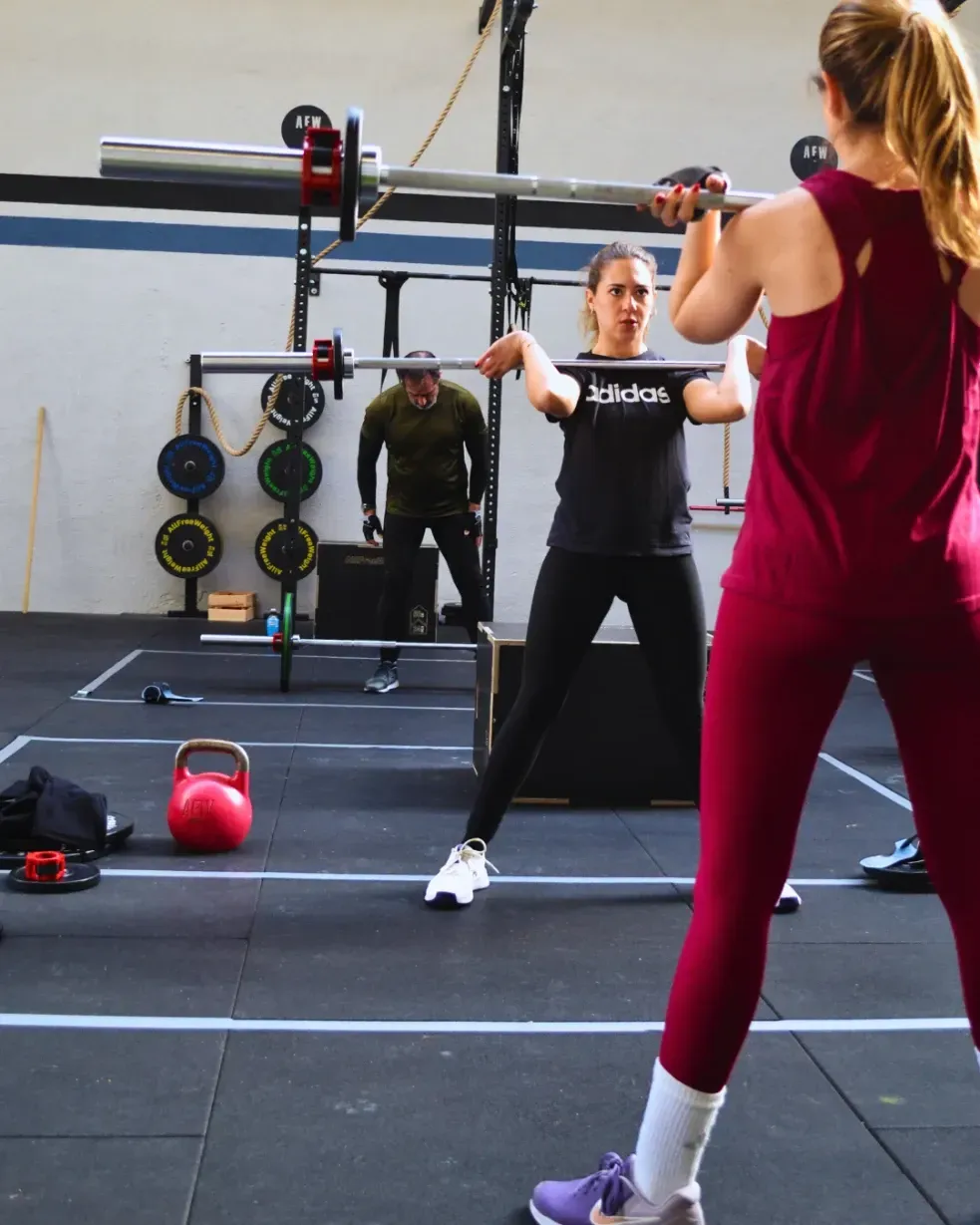 Gente levantando pesas en un gimnasio. Una mujer con camiseta negra, otra con camiseta roja. Otros al fondo.