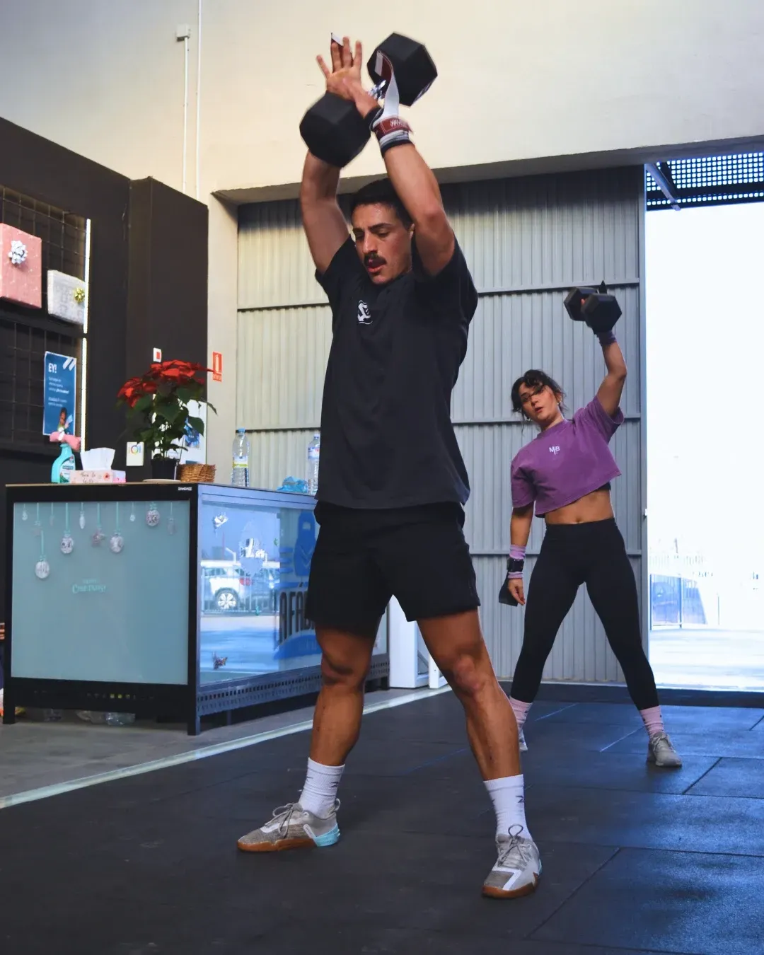 Dos personas ejercitándose con pesas rusas en un gimnasio. El hombre está arriba. Ambos visten ropa deportiva.