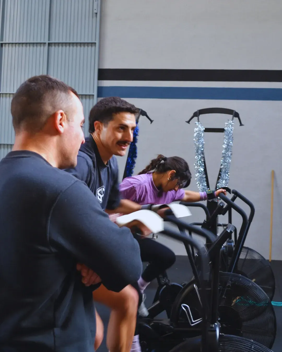 Personas en un gimnasio: dos hombres observan a una persona en una bicicleta estática.