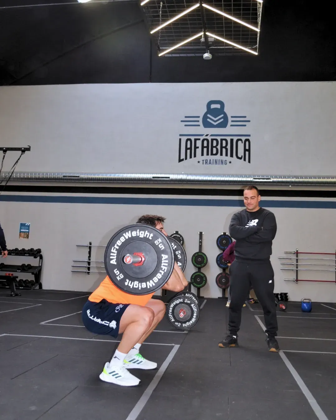 Hombre en cuclillas con barra, observado por un entrenador, dentro del gimnasio 
