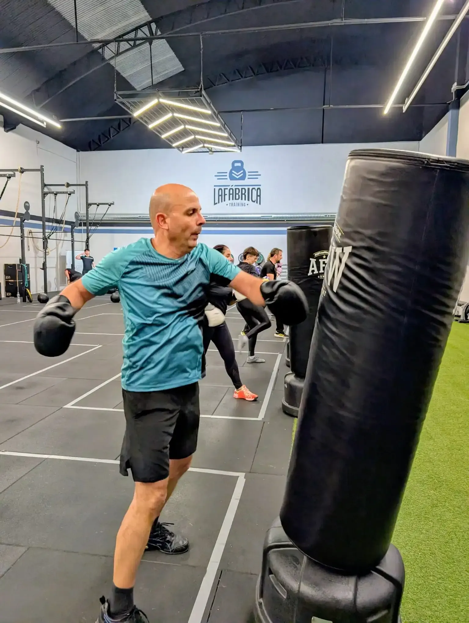 Un hombre boxea con un saco pesado en un gimnasio. Otras personas también entrenan.