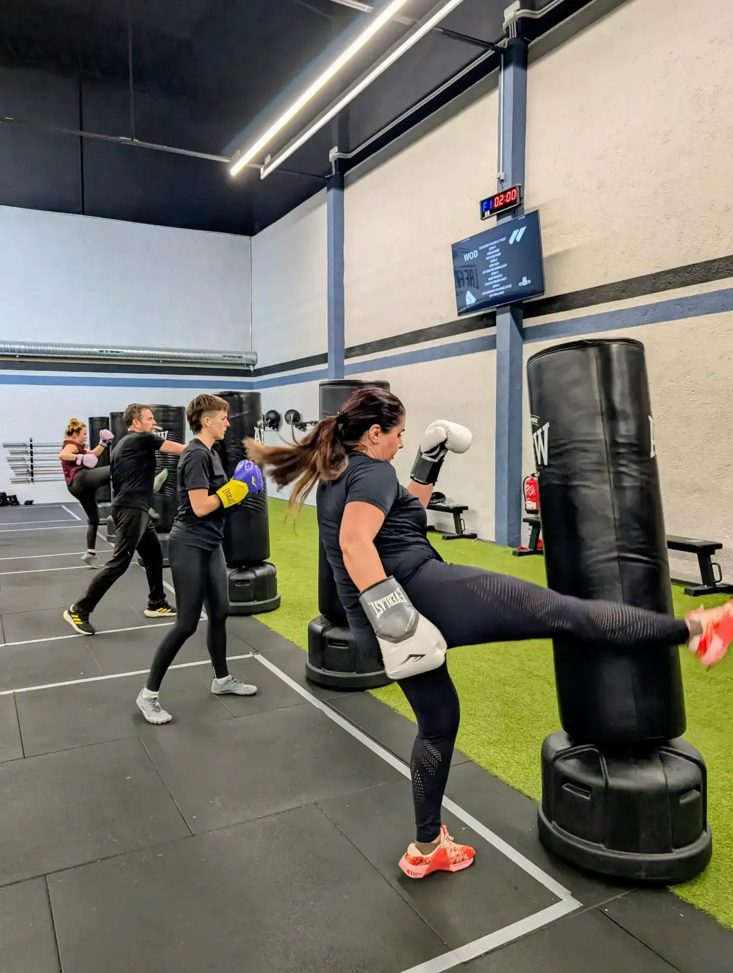 Un hombre salta a una caja en un gimnasio, vigilado por un perro. Al fondo, otra persona levanta pesas.