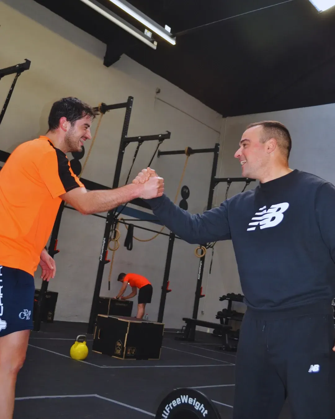 Dos hombres chocan los puños en un gimnasio, otra persona salta sobre una caja en el fondo.