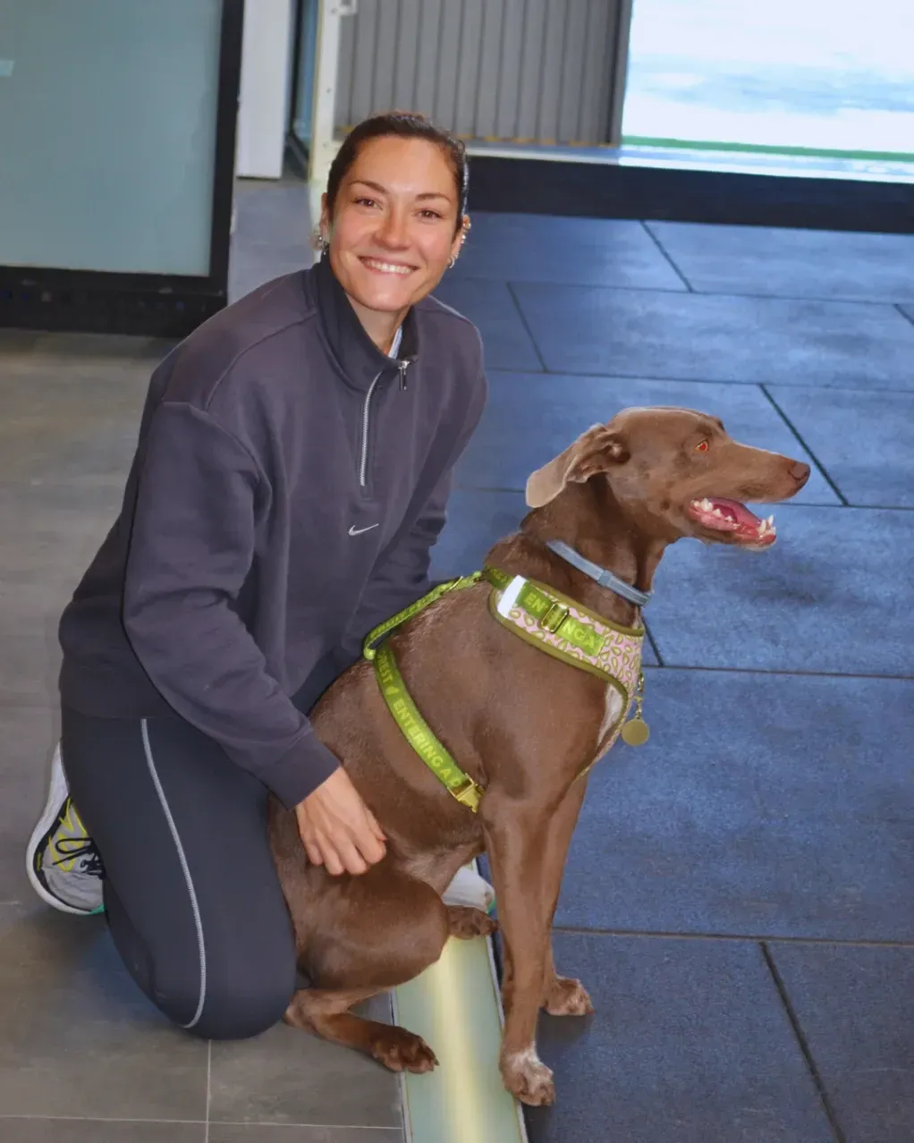 Una mujer arrodillada acaricia a un perro marrón con arnés. Están en un interior sobre un suelo gris.