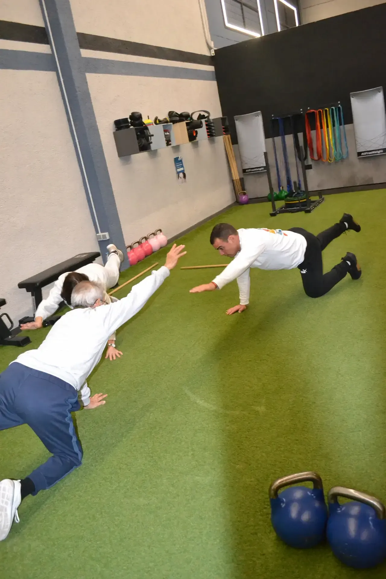 Dos personas ejercitándose en el suelo verde del gimnasio, una estirando los brazos y la otra en plancha.