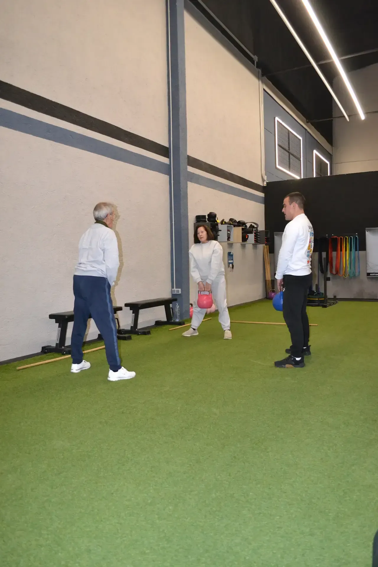 Tres personas en un gimnasio; una mujer sostiene una pesa rusa rosa, dos hombres observan. Suelo de césped verde.