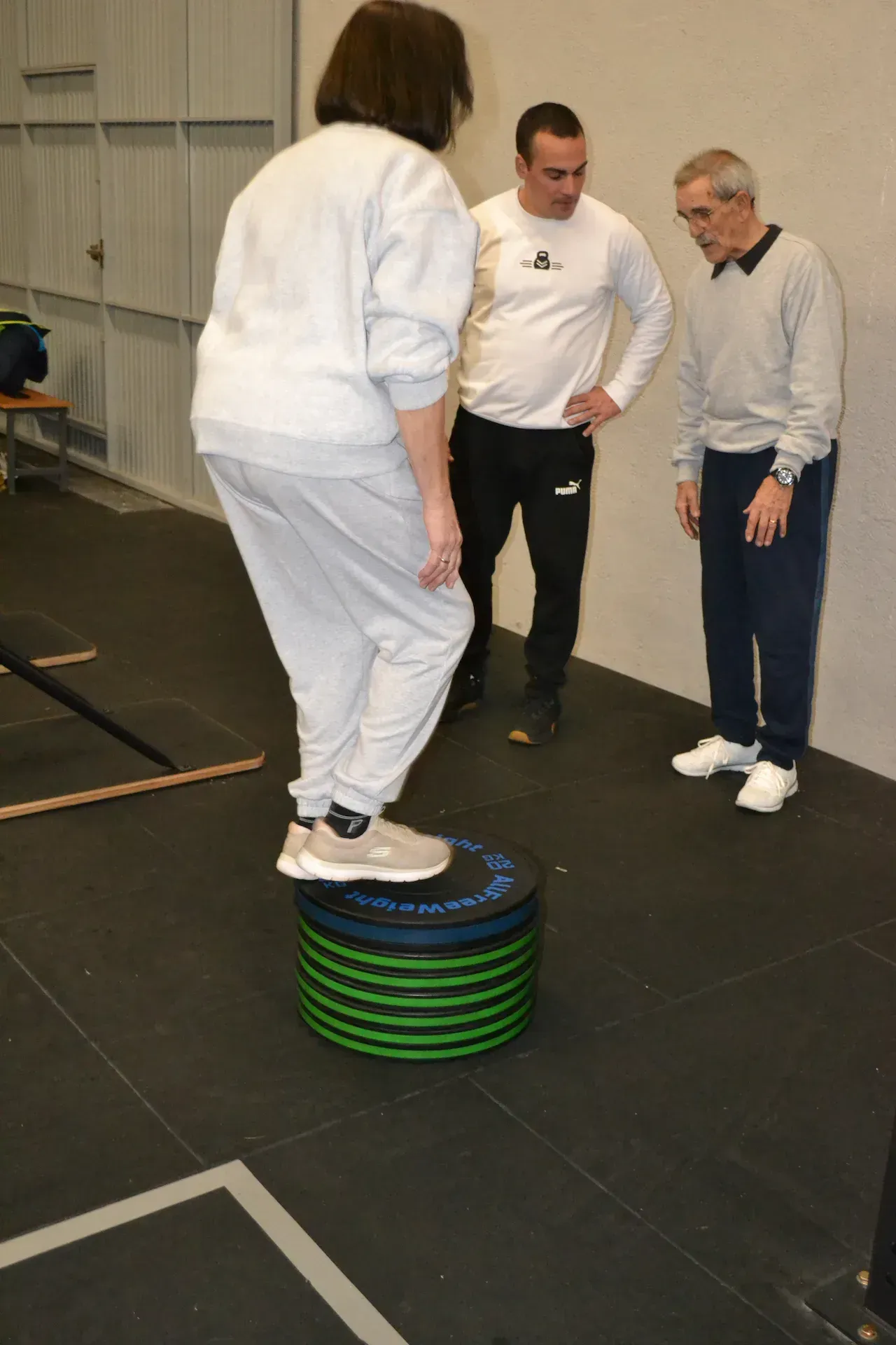 Mujer haciendo equilibrio sobre una plataforma apilada, mientras dos hombres observan en un gimnasio.