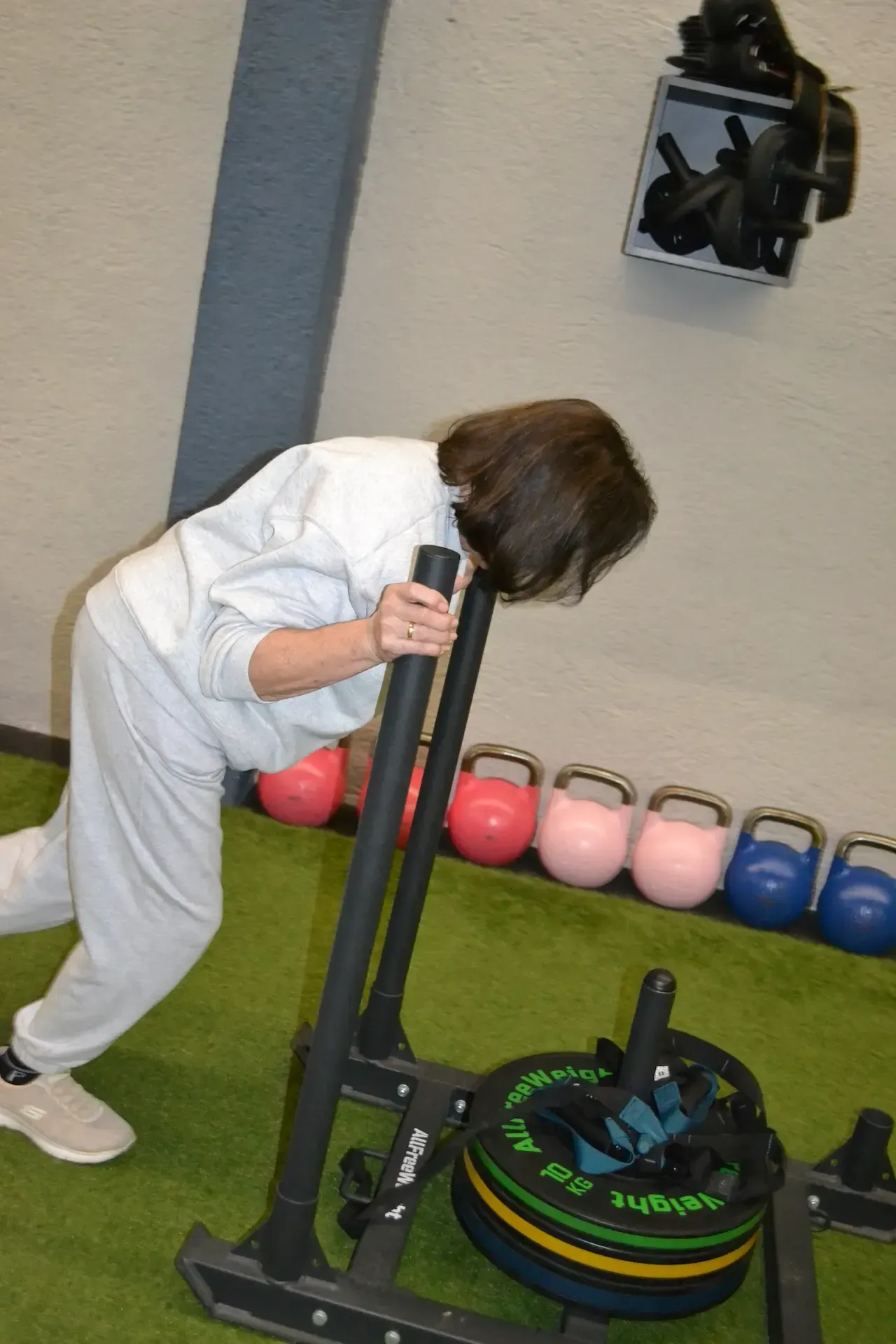 Mujer empujando un trineo con peso sobre un suelo de césped verde en un gimnasio, agarrándose de las manijas.