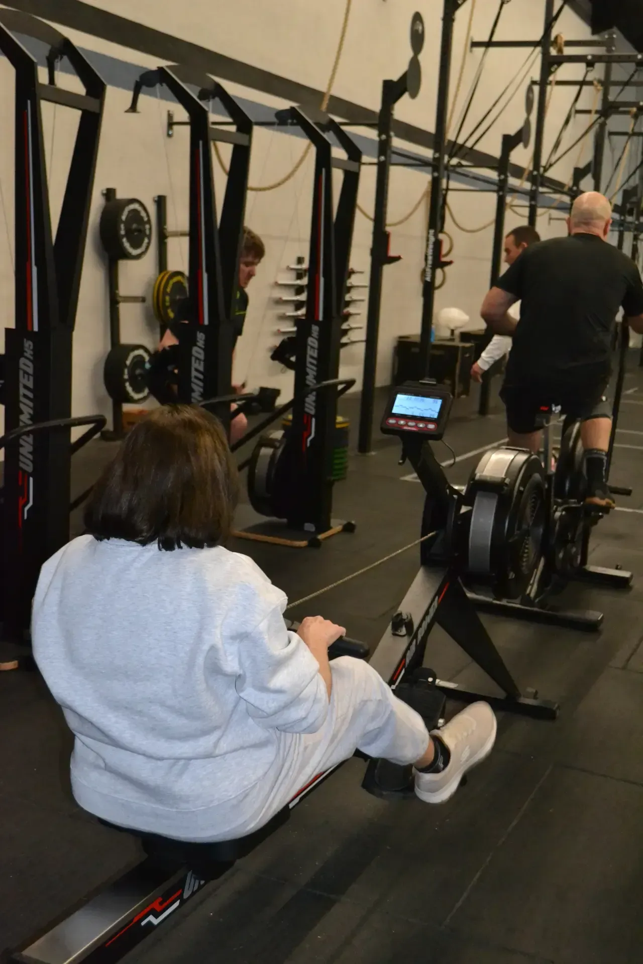 Gente levantando pesas en un gimnasio. Barras, cajas y una persona con camisa blanca y mallas marrones.