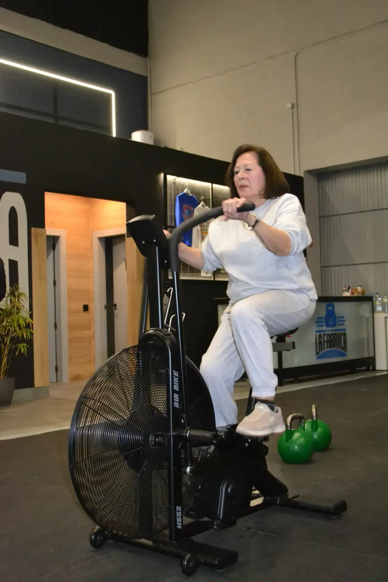 Mujer haciendo ejercicio en una bicicleta estática negra en un gimnasio, con un chándal gris. Dos pesas rusas verdes cerca.