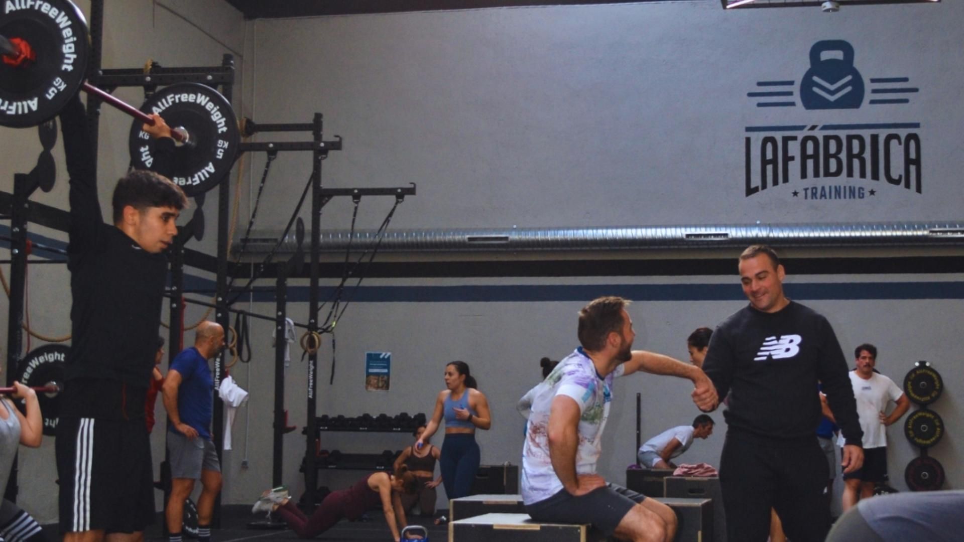 Gente en un gimnasio: una persona levanta una barra, un entrenador observa, otros hacen ejercicio.