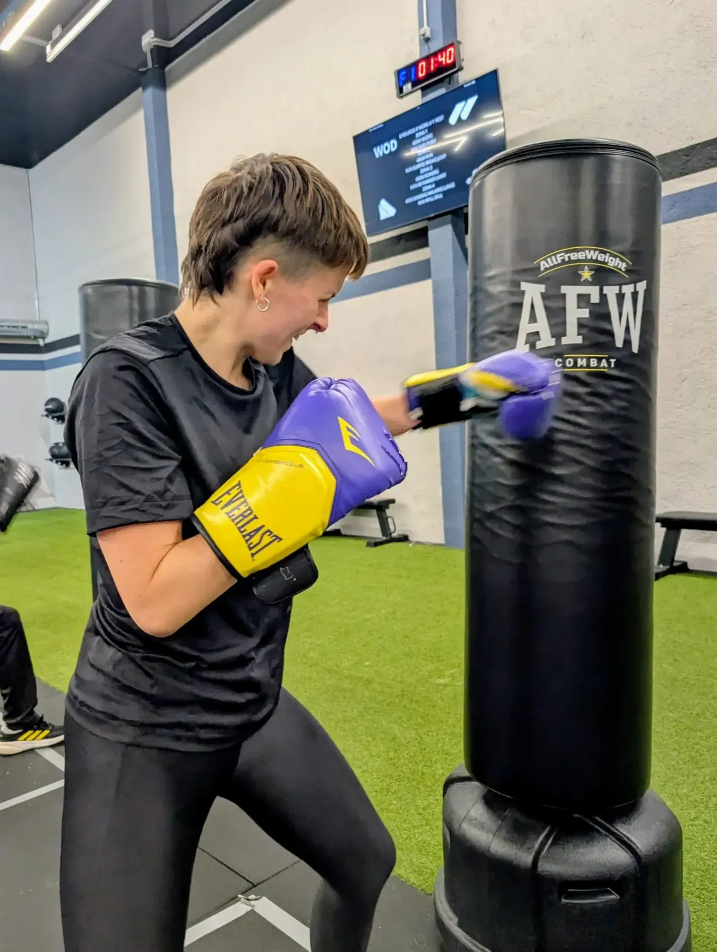 Una persona con guantes golpea un saco de boxeo negro en un gimnasio. Lleva ropa negra y el pelo corto.