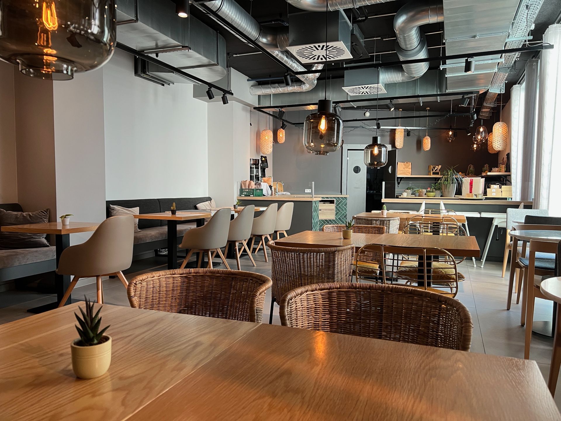 Ein hell erleuchtetes Restaurantinterieur mit Tischen, Korbstühlen und einem Barbereich im Industriestil mit offener Decke.