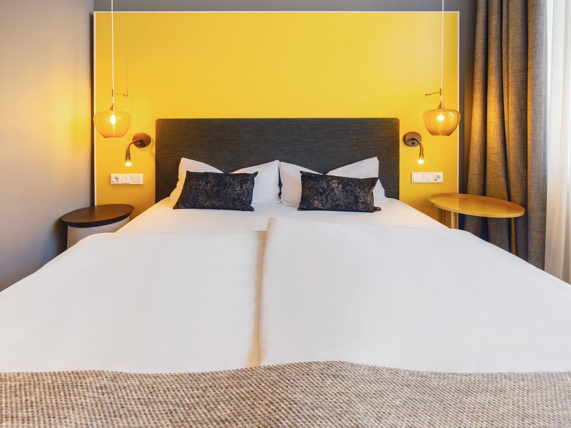 Ein modernes Hotelzimmer mit einer leuchtend gelben Akzentwand, einem dunkelgrauen Kopfteil, weißer Bettwäsche und zwei Hängelampen.
