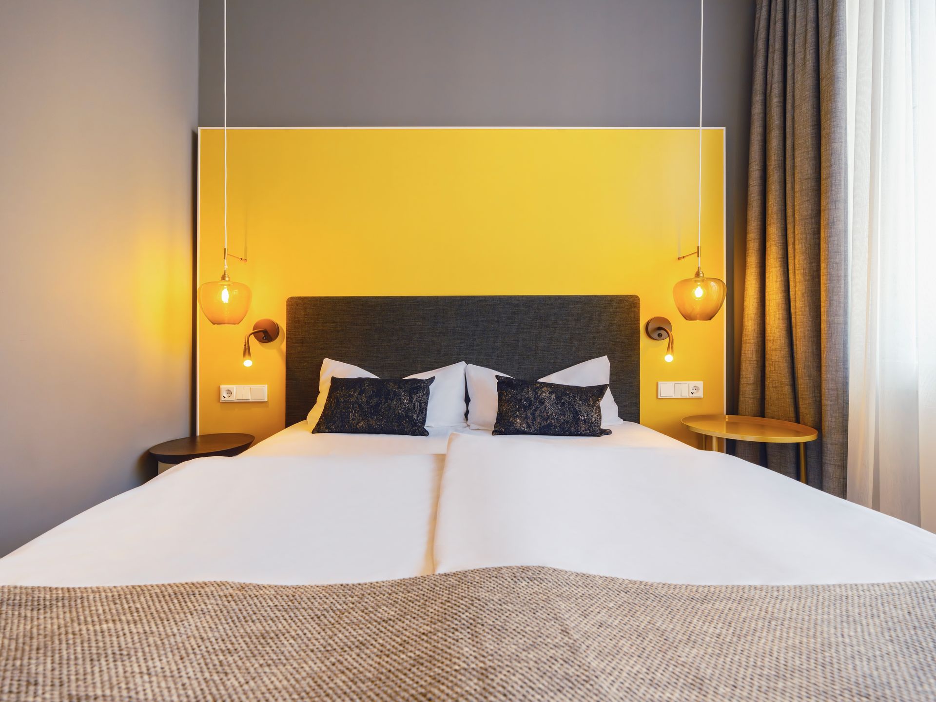 Ein modernes Hotelbett mit weißer Bettwäsche, dunklen Kissen und einem leuchtend gelben Kopfteil mit hängenden Pendelleuchten.