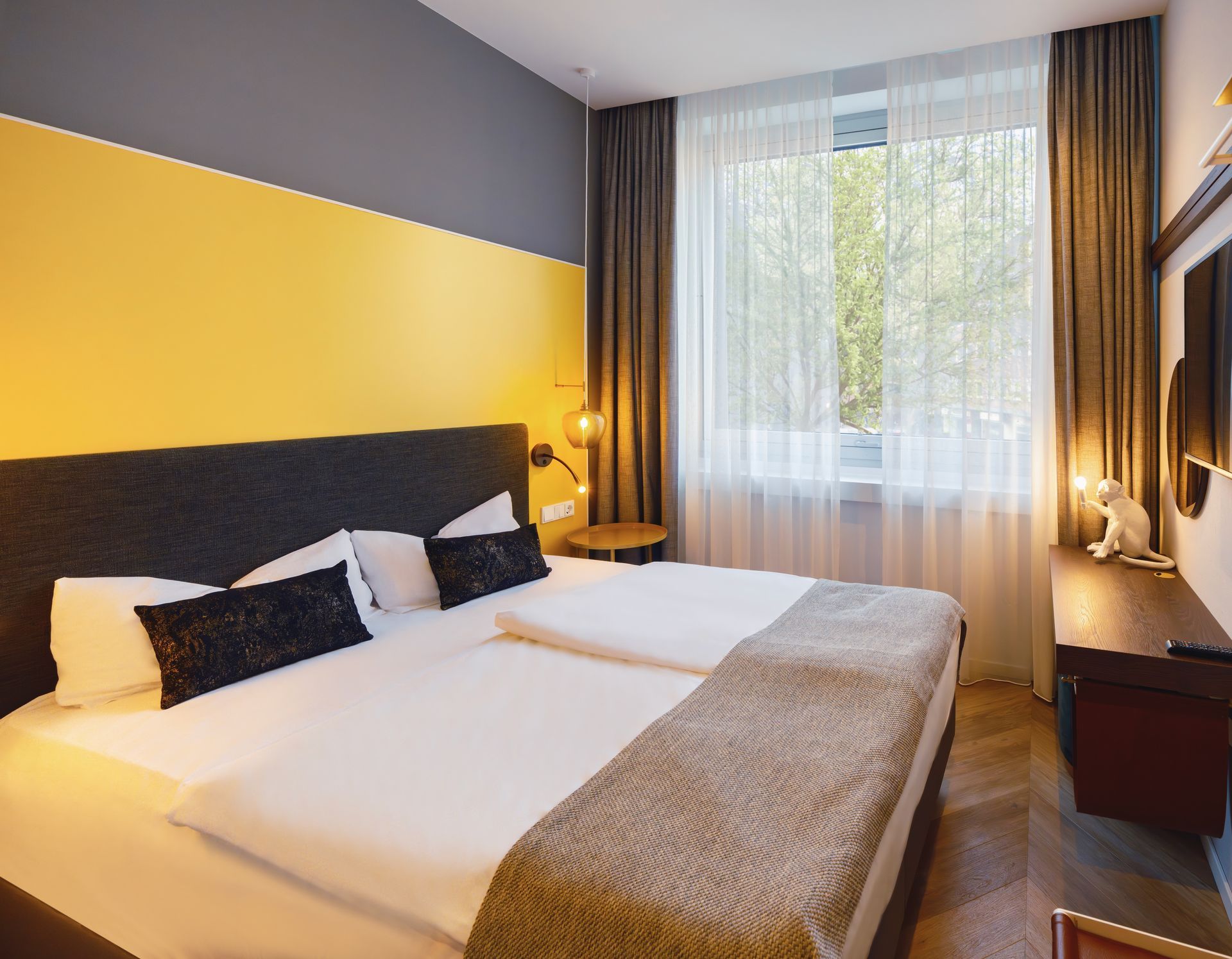 A modern hotel room with two single beds, a yellow accent wall, and a large window with sheer curtains