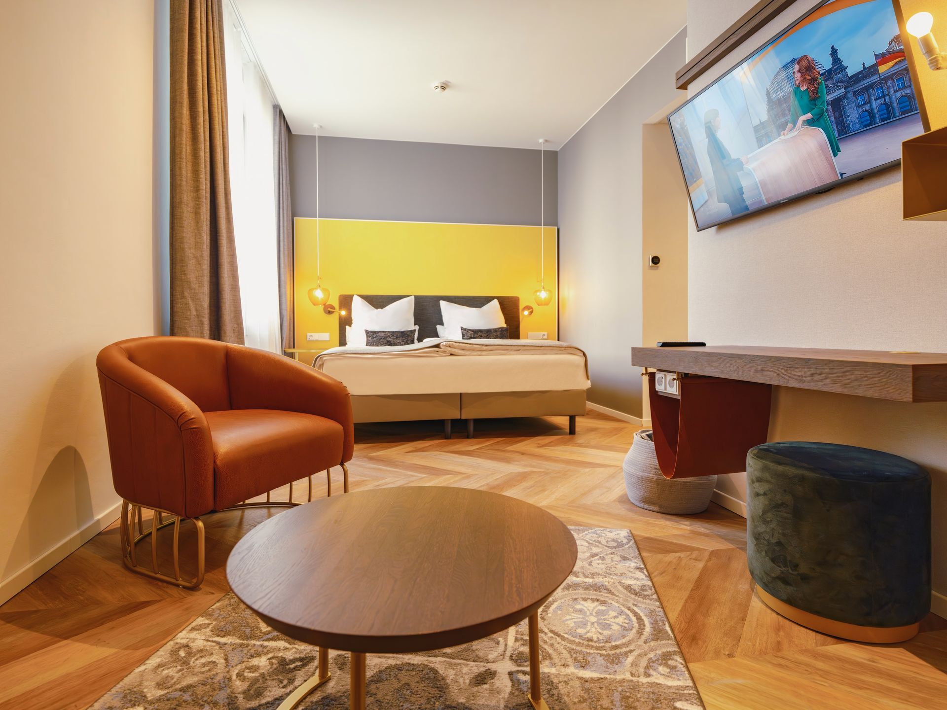 Ein modernes Hotelzimmer mit einem Bett vor einer gelben Wand, einem braunen Sessel, einem runden Couchtisch und einem an der Wand montierten Fernseher.