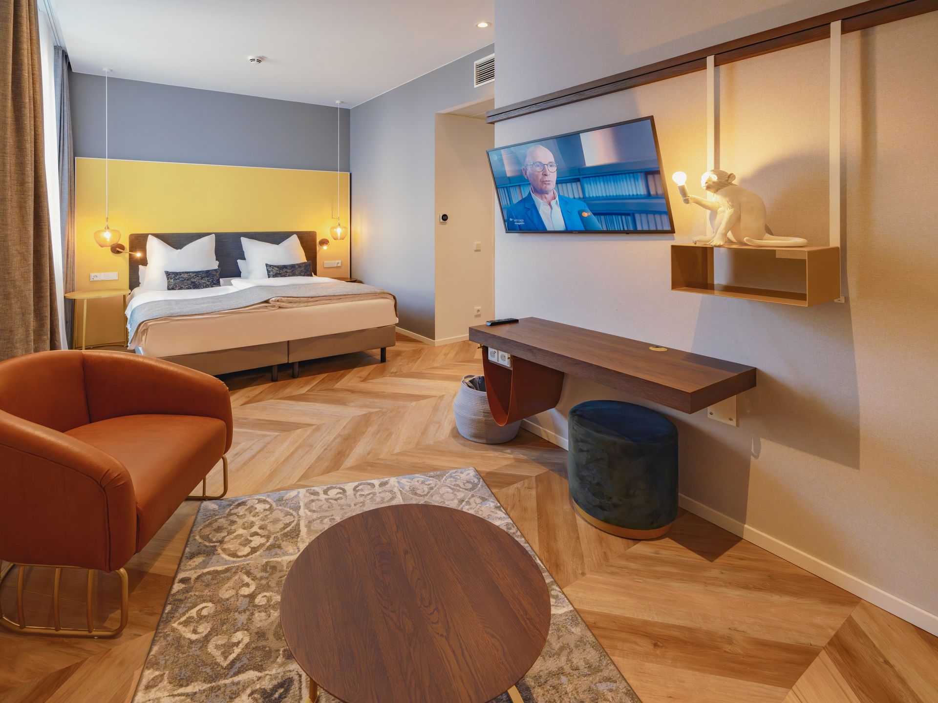 Ein modernes Hotelzimmer mit einem Bett vor einer gelben Wand, einem orangefarbenen Sessel, einem Couchtisch und einem an der Wand montierten Fernseher.