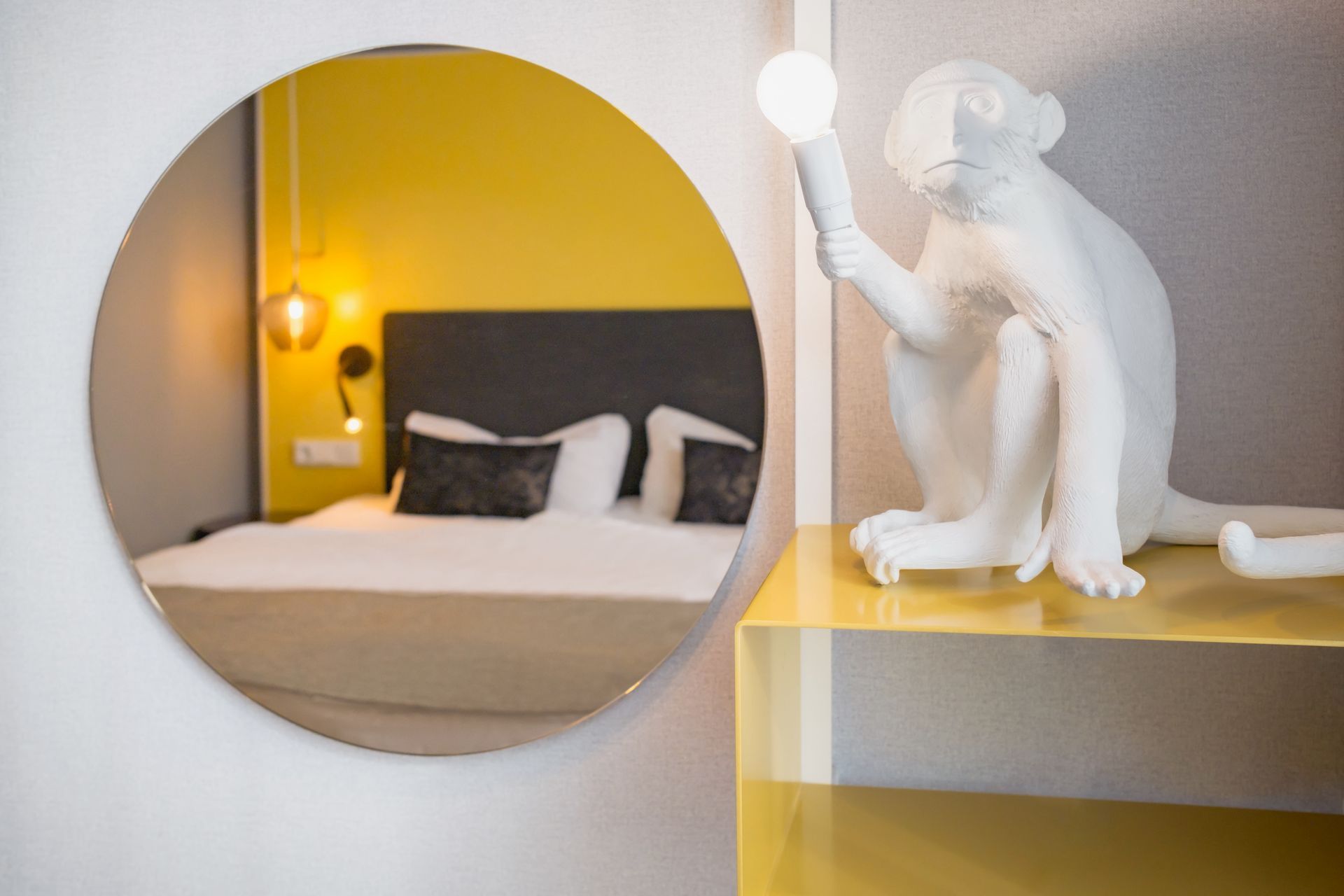 Ein runder Spiegel reflektiert ein Schlafzimmer mit Bett und gelber Wand, daneben steht eine weiße, affenförmige Lampe auf einem gelben Regal.