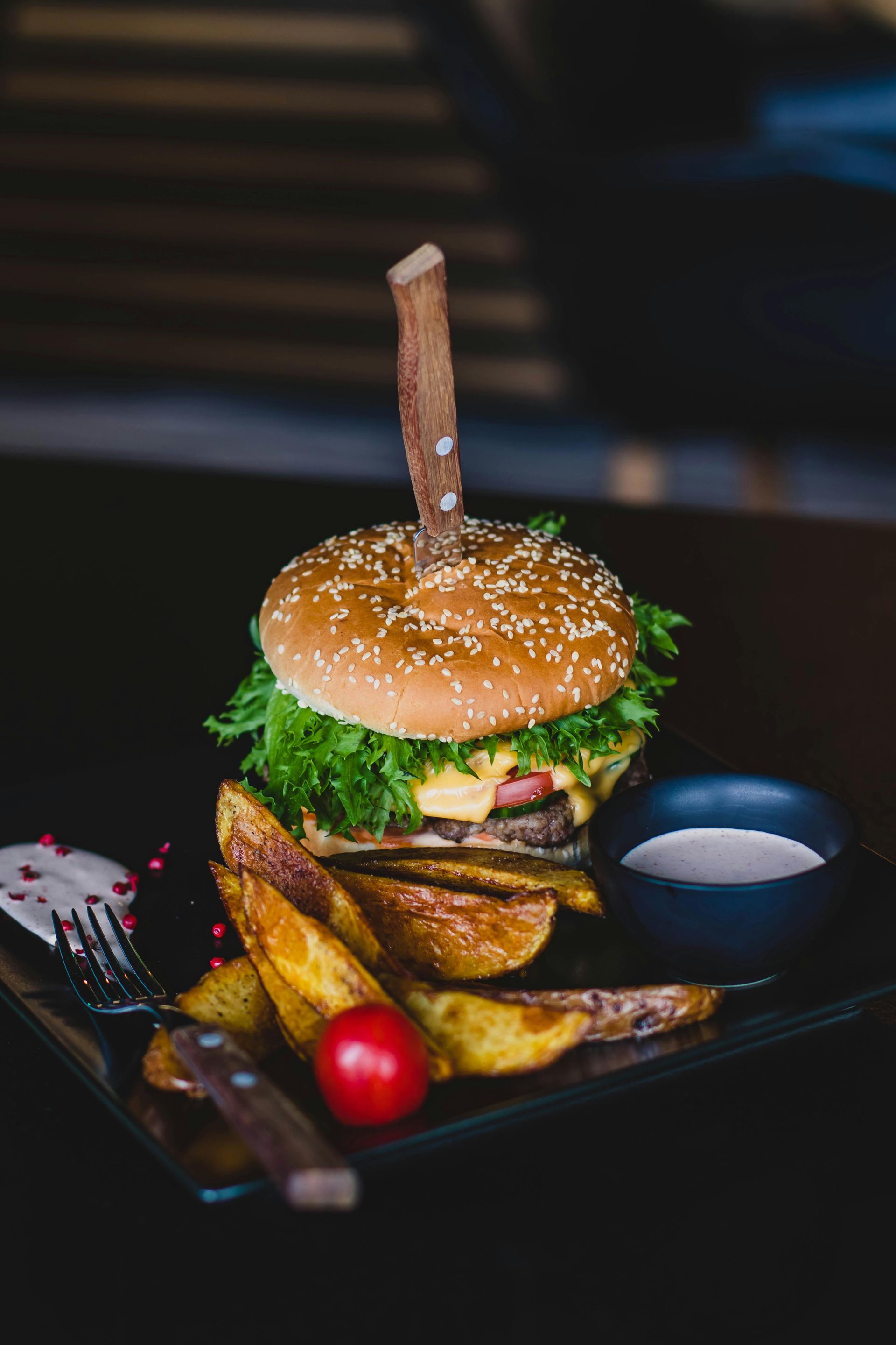 Gourmet-Burger mit Pommes frites und Dip-Sauce auf einem schwarzen Teller, garniert mit einem Holzspieß und einem Sesambrötchen.