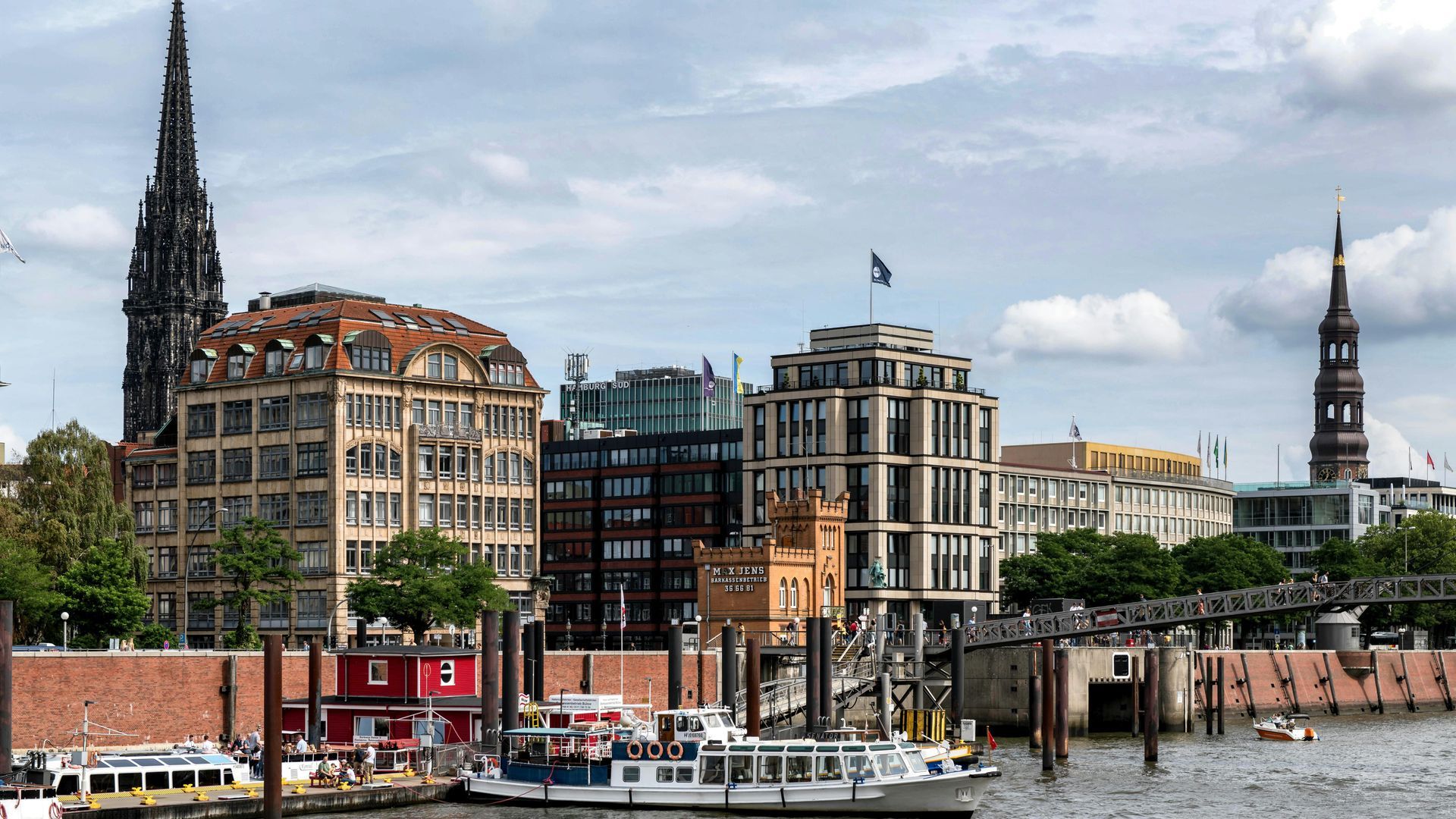 Ein Blick auf die Uferpromenade von Hamburg, Deutschland, mit historischen Kirchtürmen, traditionellen Backsteingebäuden und Ausflugsbooten.