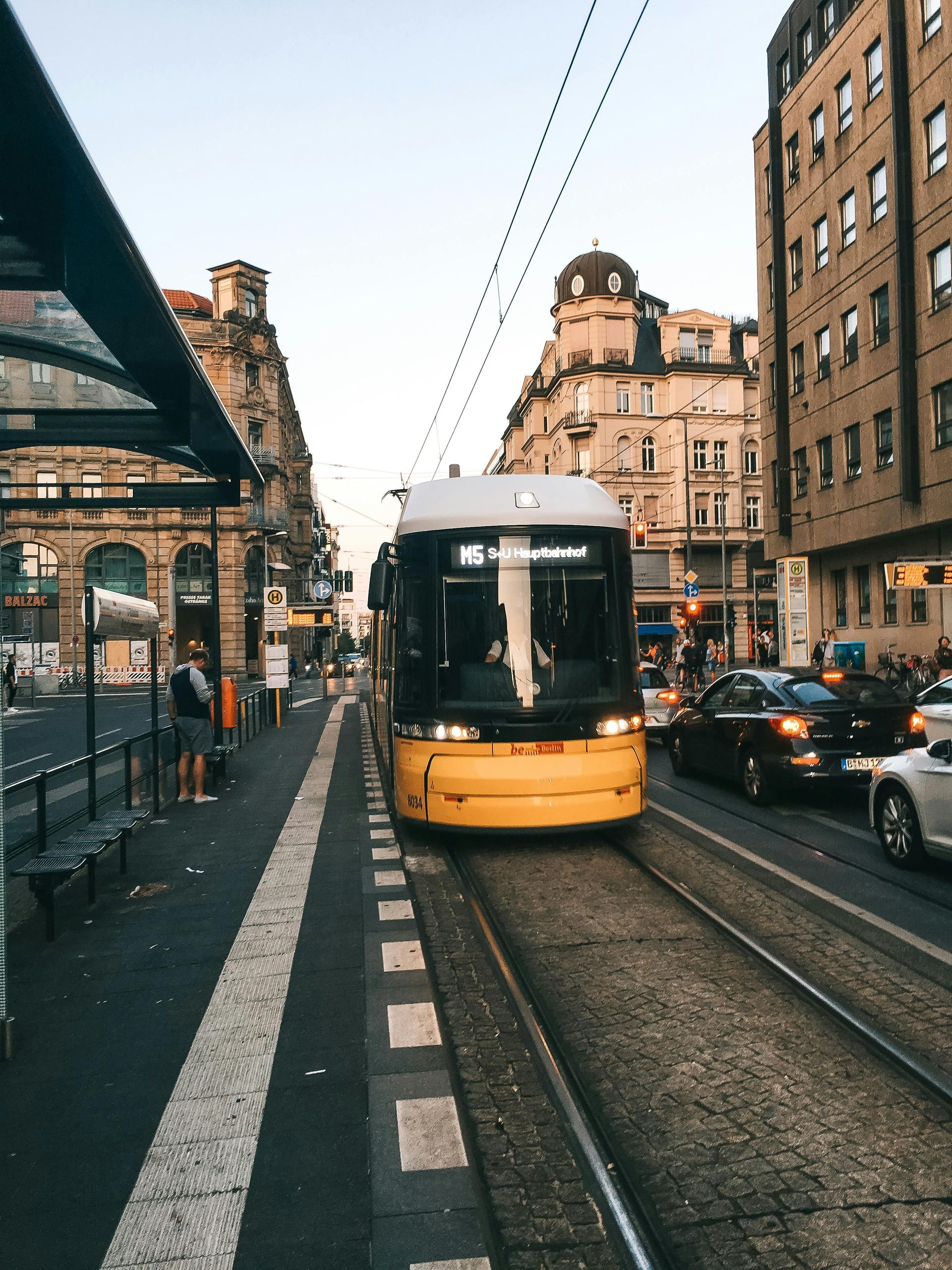 Eine gelbe Straßenbahn nähert sich tagsüber einem Stadtbahnhof, umgeben von Autos und prunkvollen Gebäuden.