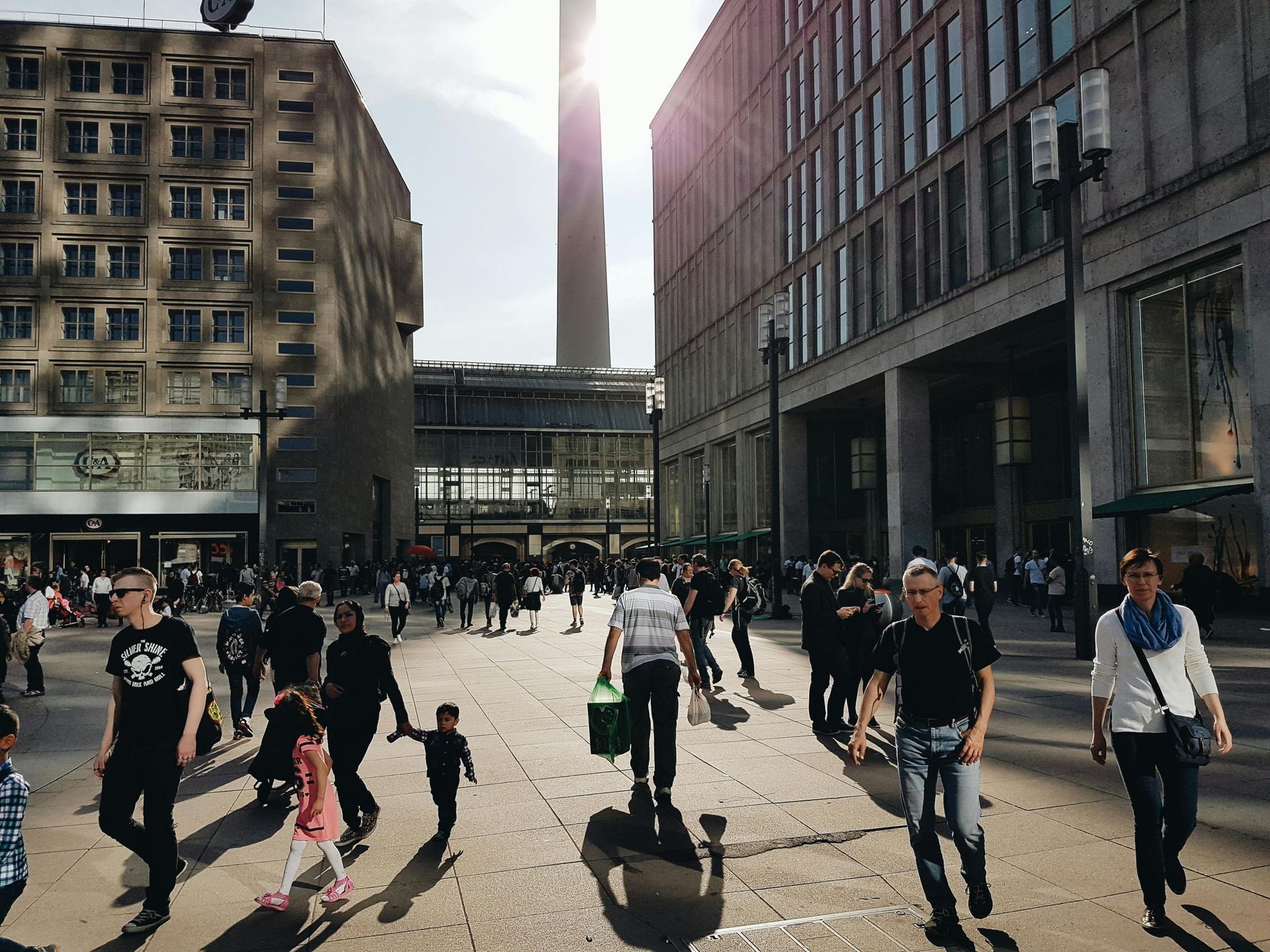 Menschen spazieren über einen sonnigen, gepflasterten Platz in einer Stadt mit großen, modernen Gebäuden und einem hohen Turm in der Ferne.