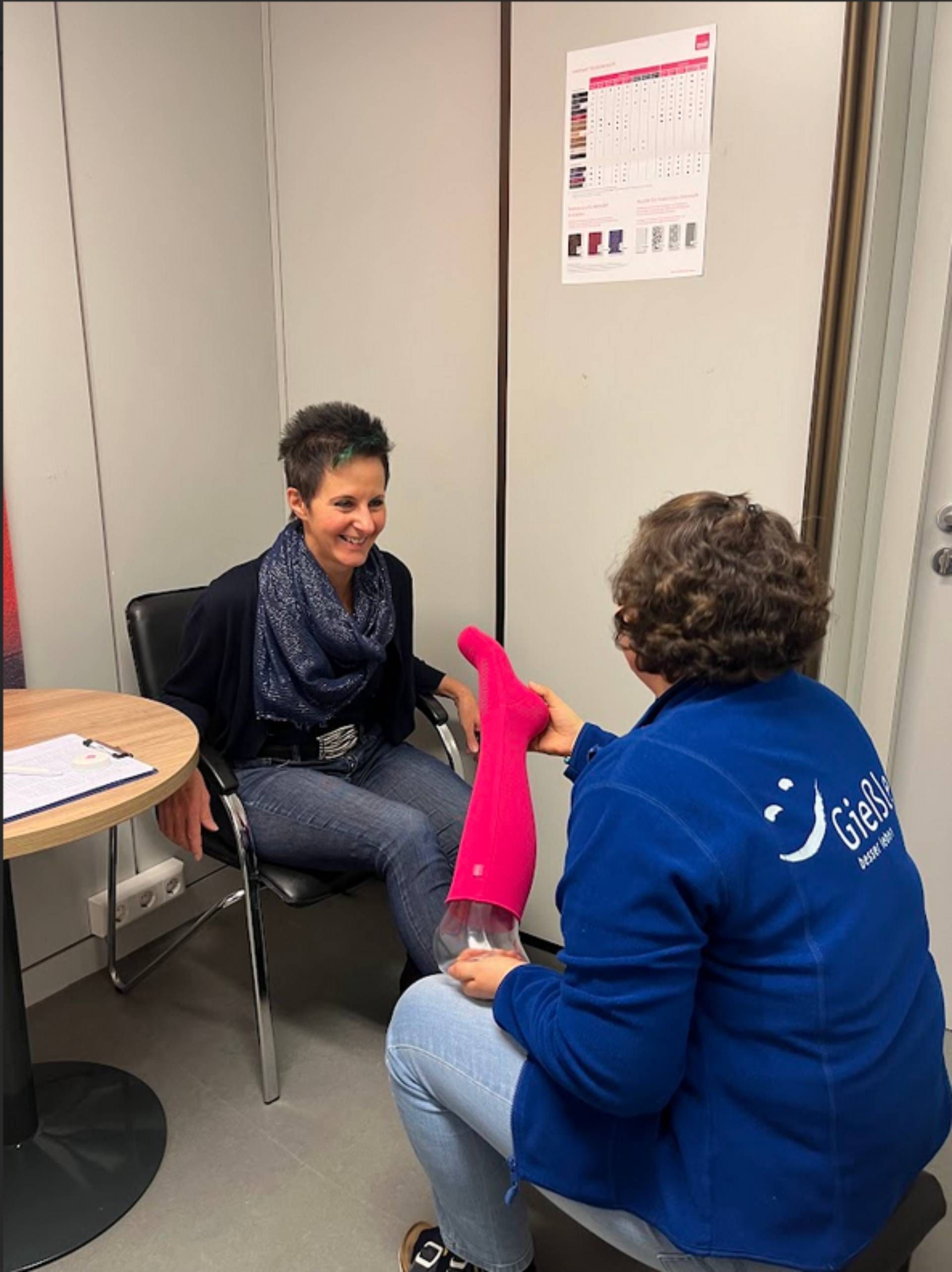 Zwei Personen in einem Raum, eine sitzt auf einem Stuhl, die andere hält ein pinkes Kompressionsstrumpf