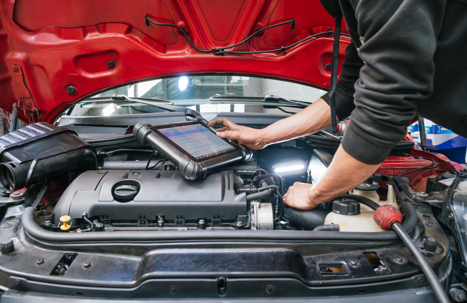 Un mécanicien utilise une tablette de diagnostic sous le capot ouvert d’une voiture rouge, dans un garage automobile.