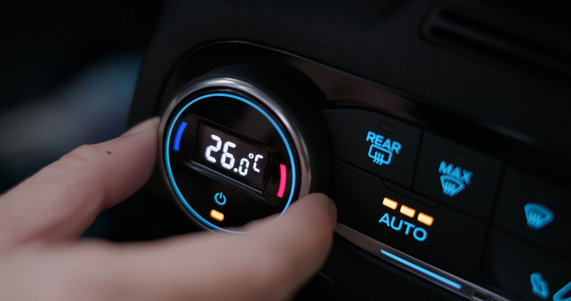 Une main règle le bouton de climatisation d’une voiture, avec un affichage de température rétroéclairé en bleu et des boutons de climatisation.
