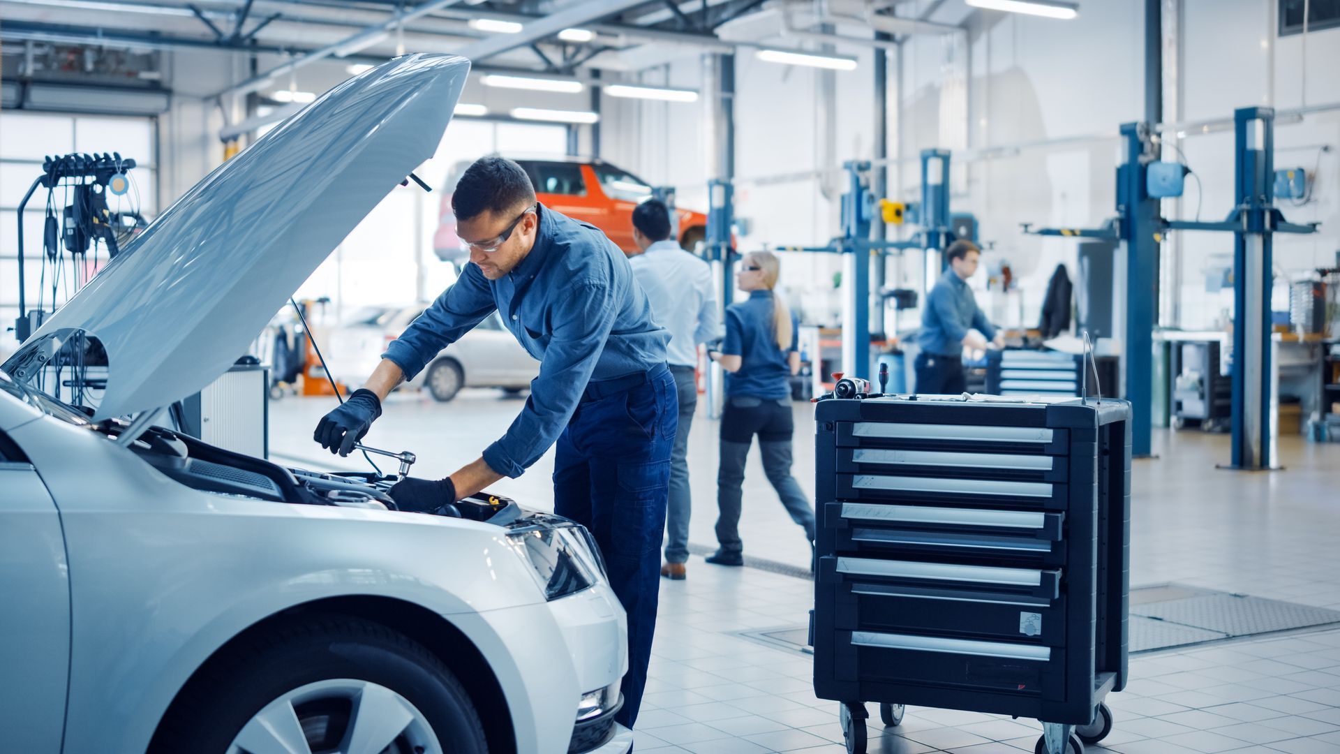 Mécanicien inspectant le moteur d’une voiture dans un atelier de réparation automobile lumineux, avec une servante à outils à proximité.