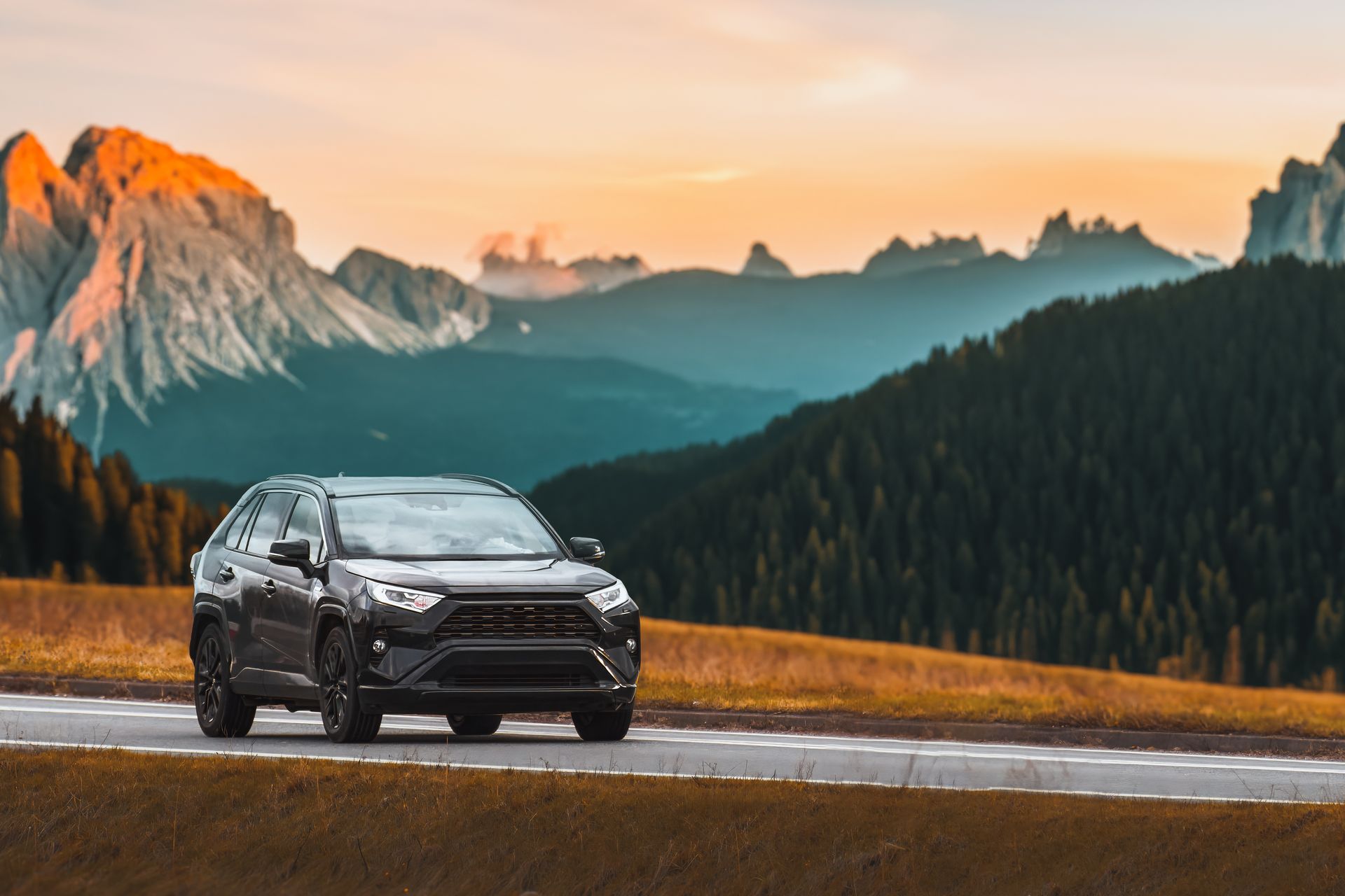 Un SUV noir roule sur une route de montagne au coucher du soleil, avec des sommets enneigés en arrière-plan.