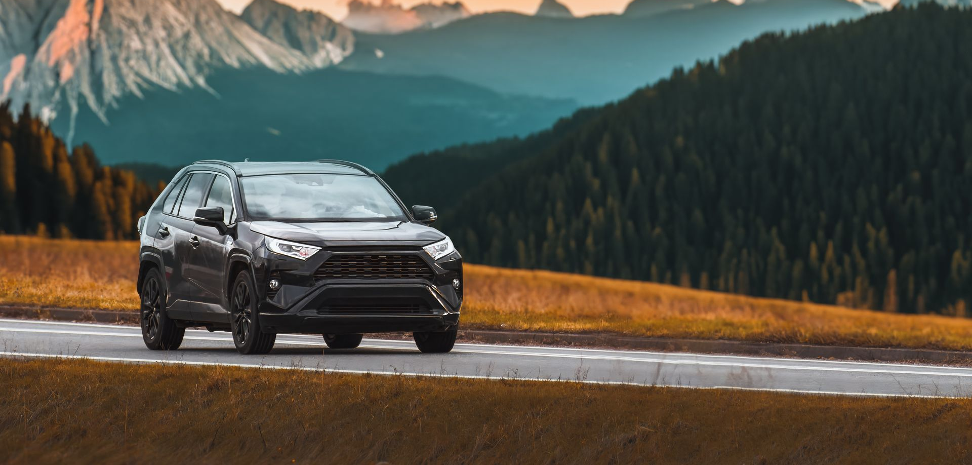 Un SUV noir roule sur une route de montagne, avec une forêt et des sommets enneigés en arrière-plan.
