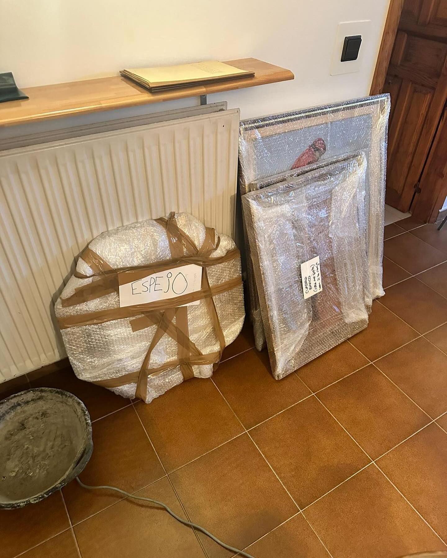 Dos cuadros envueltos en plástico se encuentran sobre un suelo de baldosas junto a un radiador.